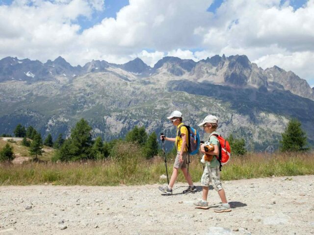 Zwei Personen, ein Erwachsener und ein Kind, tragen Hüte und Rucksäcke und gehen mit Trekkingstöcken einen Feldweg entlang. Im Hintergrund sind Berge und Bäume unter einem teilweise bewölkten Himmel zu sehen.