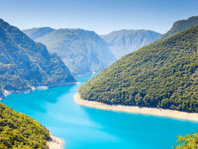 Ein leuchtend blauer Fluss schlängelt sich unter einem strahlend blauen Himmel durch eine üppige, grüne Berglandschaft mit steilen, bewaldeten Hängen zu beiden Seiten.