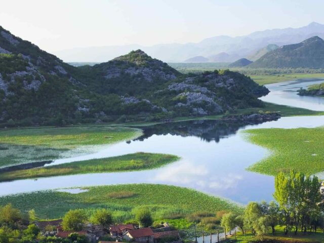 Ein malerischer Fluss schlängelt sich durch grüne Hügel und Feuchtgebiete, in denen sich der Himmel spiegelt. Die Berge verschwinden in der Ferne, und im Vordergrund sind zwischen den Bäumen Häuser mit roten Dächern zu sehen.