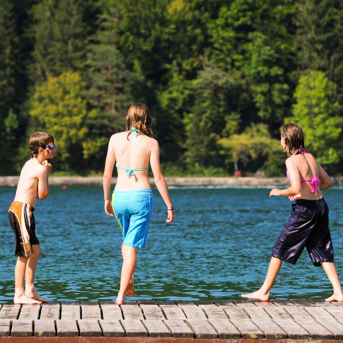 Drei Kinder in Badeanzügen und Badehosen stehen auf einem Holzsteg an einem See und bereiten sich darauf vor, ins Wasser zu gehen. Der Hintergrund ist mit grünen Bäumen und hellem Sonnenlicht gefüllt.