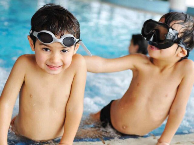 Zwei Jungen mit Schwimmbrillen lächeln und spielen am Rande eines Hallenbades. Ein Junge steht im Wasser und legt dem anderen die Hand auf die Schulter, während sich der andere Junge am Beckenrand festhält.