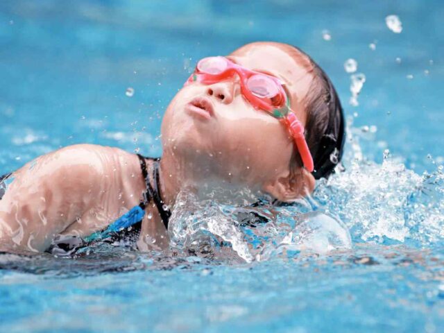 Ein kleines Kind mit rosafarbener Schwimmbrille und Badekappe schwimmt in einem Schwimmbecken und lässt das Wasser um sich herum spritzen.