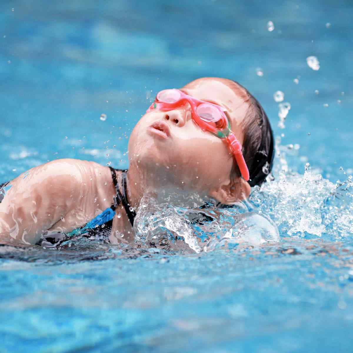Ein kleines Kind mit rosafarbener Schwimmbrille und Badekappe schwimmt in einem Schwimmbecken und lässt das Wasser um sich herum spritzen.
