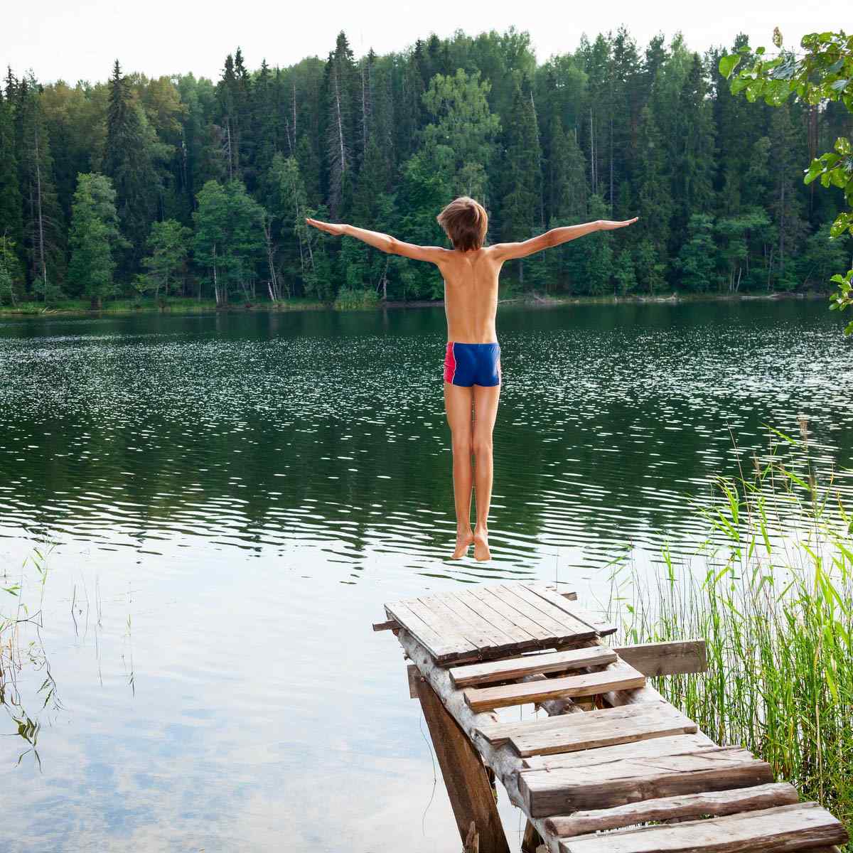 Ein Kind steht am Rande eines hölzernen Stegs mit ausgestreckten Armen und bereitet sich darauf vor, in einen ruhigen See zu springen, der von einem dichten grünen Wald umgeben ist.