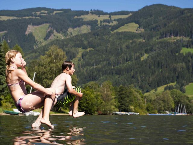 Zwei Kinder in Badeanzügen springen an einem sonnigen Tag gemeinsam in einen See und planschen im Wasser, mit baumbestandenen Hügeln und Booten im Hintergrund.