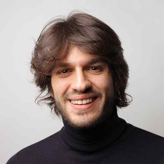 Ein Mann mit mittellangem braunem Haar und Bart lächelt in die Kamera. Er trägt einen schwarzen Rollkragenpullover vor einem schlichten hellgrauen Hintergrund.