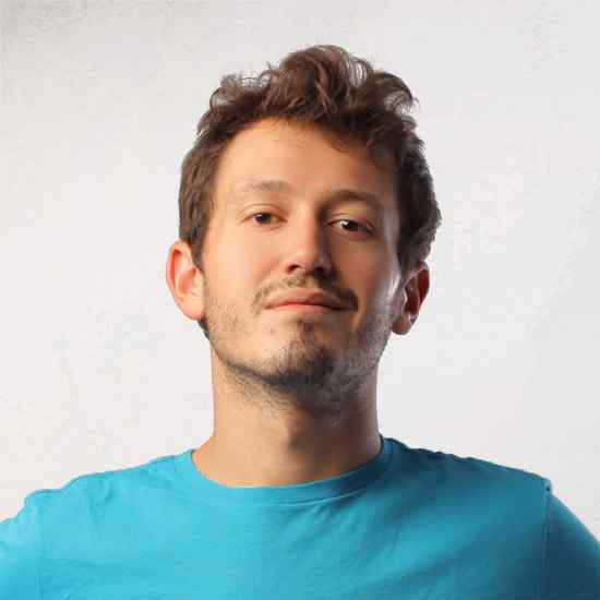 Ein junger Mann mit kurzen braunen Haaren und heller Gesichtsbehaarung trägt ein hellblaues T-Shirt und blickt selbstbewusst in die Kamera vor einem schlichten hellen Hintergrund.