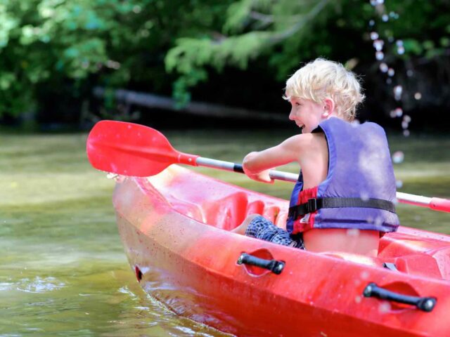 Ein kleiner Junge mit Schwimmweste paddelt in einem roten Kajak auf einem ruhigen, flachen Fluss, der von grünen Bäumen umgeben ist.