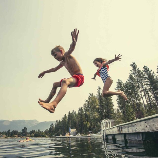 Zwei Kinder in Badeanzügen springen von einem Steg in einen See, Arme und Beine in der Luft ausgestreckt, mit Bäumen und Bergen im Hintergrund an einem sonnigen Tag.