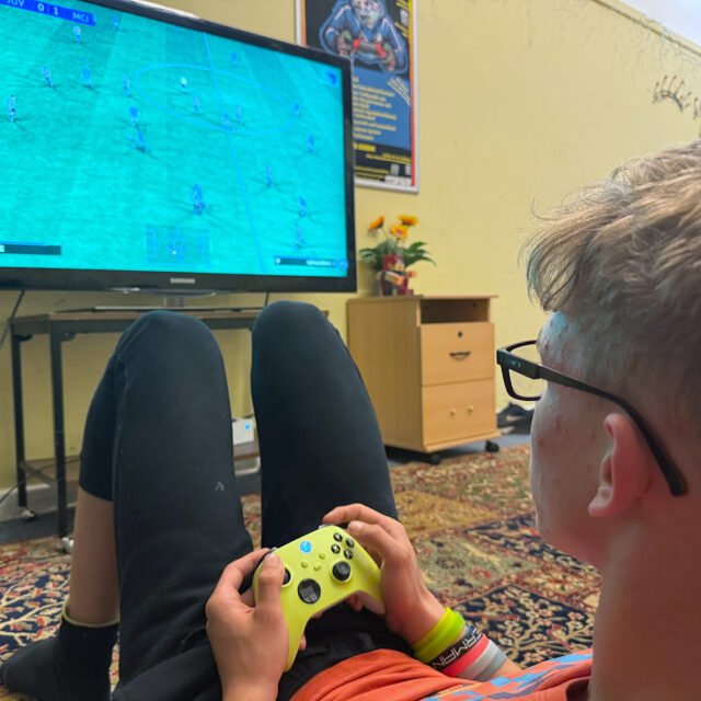 Eine Person mit Brille sitzt auf dem Boden, hält einen gelben Game-Controller in der Hand und spielt ein Fußball-Videospiel auf einem großen Fernseher in einem gemütlichen Raum mit gemustertem Teppich und Möbeln.