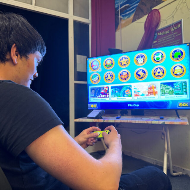 Eine Person sitzt auf einem Stuhl und spielt ein Videospiel mit einem Controller. Auf dem Fernsehbildschirm davor ist ein buntes Spielmenü mit mehreren runden Symbolen zu sehen. Der Raum hat einen Schreibtisch, Poster und ein Fenster im Hintergrund.