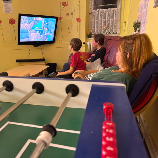 Drei Personen sitzen auf einer Couch und schauen auf einen Fernsehbildschirm, der ein Videospiel zeigt. Im Vordergrund steht ein Tischfußballtisch mit roten Kugeln. Der Raum ist gelb gestrichen und an den Wänden mit Blumen verziert.