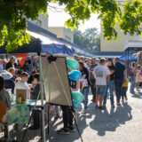 Ein lebhafter Jahrmarkt im Freien mit vielen Menschen, Marktständen und bunten Dekorationen. Einige Menschen sitzen an Tischen, andere gehen zwischen den Ständen umher. Luftballons und festliche Wimpel sind zu sehen, und das Sonnenlicht fällt durch die grünen Blätter der Bäume.