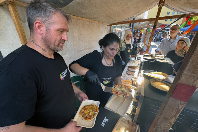 Zwei Personen servieren persönliche Pizzen an einem Imbissstand im Freien und reichen den Kunden Teller. Andere Menschen warten in einer Schlange unter einem Zelt, während im Hintergrund bunte Fahnen und eine Menschenmenge zu sehen sind.