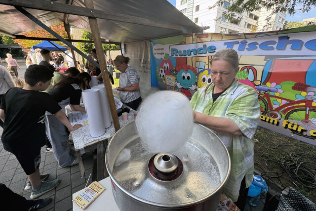 Eine Frau dreht Zuckerwatte an einem Veranstaltungsstand im Freien. In der Nähe versammeln sich Kinder und Erwachsene an Tischen unter einem Zelt. Im Hintergrund hängt ein buntes Banner mit Zeichentrickfiguren.