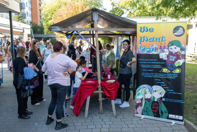 Eine Gruppe von Menschen versammelt sich bei einer Veranstaltung im Freien um einen Holzstand mit roten Tüchern. Ein buntes Banner mit Cartoon-Figuren und dem Text We Want You! steht neben dem Stand. Im Hintergrund sind Bäume und Gebäude zu sehen.