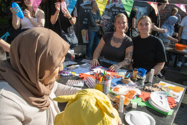 Eine Gruppe von Menschen, darunter auch Frauen und Kinder, sitzen und stehen um einen Tisch mit buntem Bastelmaterial herum und schaffen an einem sonnigen Tag Kunstwerke im Freien. Einige Personen lächeln in die Kamera.