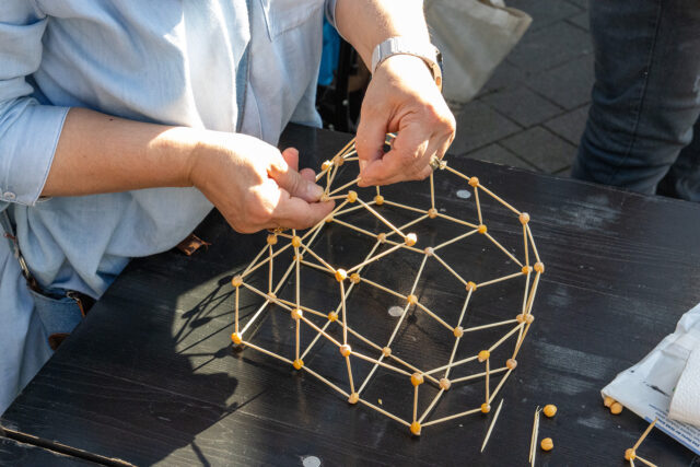 Eine Person baut eine geometrische Kuppelstruktur aus Holzstäben und kleinen runden Verbindungsstücken auf einem schwarzen Tisch zusammen.