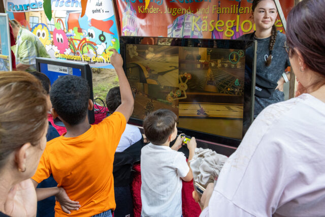 Mehrere Kinder und Erwachsene versammeln sich um einen Monitor und spielen ein Multiplayer-Videospiel bei einer Veranstaltung im Freien. Im Hintergrund sind bunte Fahnen und Dekorationen zu sehen.