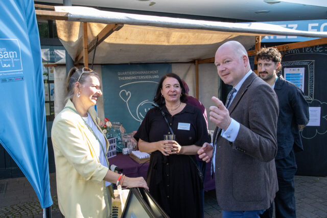 Drei Erwachsene stehen an einem Stand im Freien und unterhalten sich lächelnd, wobei ein Mann gestikuliert und zwei Frauen zuhören. Im Hintergrund sind eine Kreidetafel und Informationsbanner zu sehen.