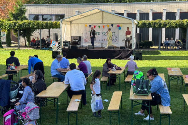 Die Menschen sitzen an Picknicktischen auf einer Wiese und sehen einem Künstler zu, der auf einer mit Wimpeln und Luftballons geschmückten Freilichtbühne auftritt. Einige Leute unterhalten sich, während Kinder in der Nähe der Tische spielen und eine Gemeinschaftsveranstaltung an einem sonnigen Tag genießen.