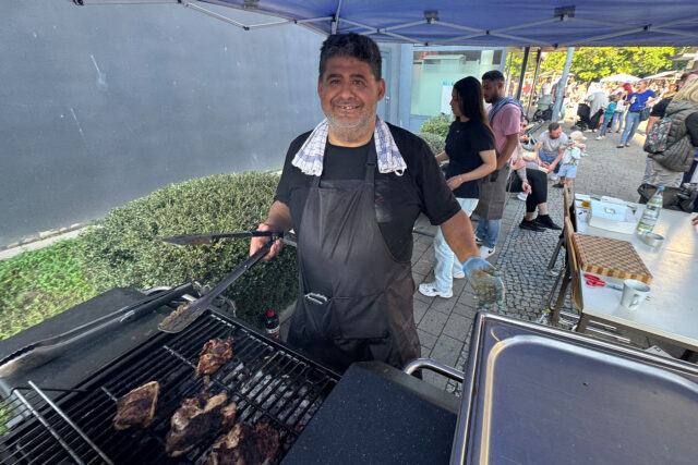 Ein Mann mit Schürze und Handschuhen grillt Fleisch bei einer Veranstaltung im Freien und lächelt in die Kamera. Im Hintergrund sind Menschen versammelt, und unter einer Überdachung sind Tische mit Lebensmitteln und Vorräten aufgebaut.