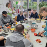 Eine Gruppe von Menschen unterschiedlichen Alters sitzt und steht um einen Tisch herum und schält und schneidet verschiedene Gemüsesorten in einem hellen, mit Luftballons dekorierten Raum mit Fenster. Schüsseln und Gemüse sind auf dem Tisch verteilt.