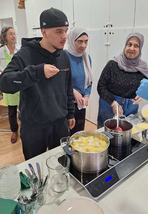 Ein junger Mann mit schwarzem Kapuzenpulli und Mütze probiert von einem Löffel neben einem Herd, während zwei Frauen in Hijabs und Schürzen lächelnd in der Nähe stehen und an einer Küchentheke mit Töpfen mit Essen kochen.