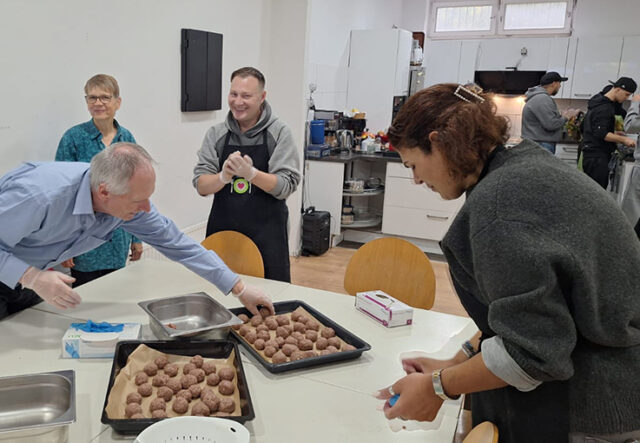 Vier Personen bereiten in einer Küche Fleischbällchen zu. Zwei Tabletts mit rohen Fleischbällchen stehen auf einem Tisch, und ein Mann greift danach, um sie anzuordnen. Die anderen lächeln und unterhalten sich; die Küche ist hell und aufgeräumt.
