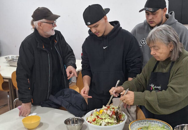 Vier Personen stehen um einen Tisch und bereiten eine große Schüssel Salat zu. Eine ältere Frau schwenkt den Salat, während zwei junge Männer und ein älterer Mann zusehen und helfen. Verschiedene Schüsseln und Zutaten stehen auf dem Tisch.