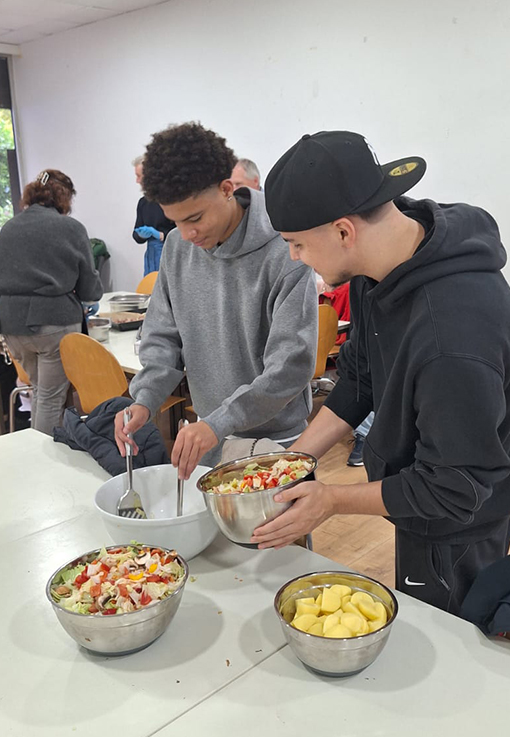 Zwei junge Männer in Kapuzenpullovern bereiten an einem Tisch das Essen zu und mischen Salate in Metallschüsseln. In einer anderen Schale liegen geschnittene Äpfel. Im Hintergrund sind weitere Personen zu sehen, einige sitzen auf Stühlen.