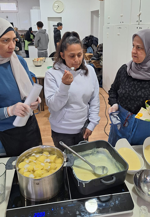 Drei Frauen stehen um einen Tisch mit Töpfen voller Essen, eine probiert von einem Löffel. Sie befinden sich in einer Küche, tragen legere Kleidung und Kopftücher, und im Hintergrund sind weitere Personen zu sehen.
