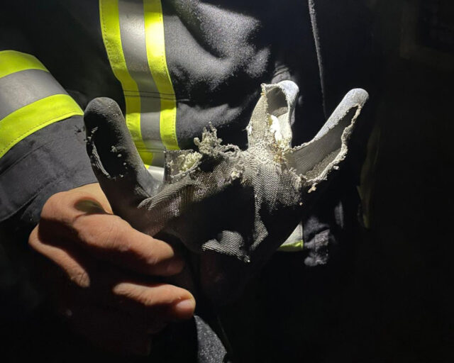 Eine Person in Feuerwehruniform hält einen stark beschädigten Handschuh mit zerrissenen Fingern und ausgefranstem Material, der von einem Lichtstrahl in einer dunklen Umgebung angestrahlt wird.