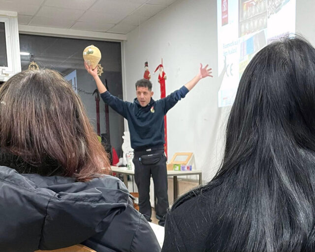 Ein Mann steht vor einem Klassenzimmer, hält mit beiden Händen ein Tamburin über dem Kopf und spricht zu einem Publikum. Zwei dunkelhaarige Personen sind von hinten im Vordergrund zu sehen.