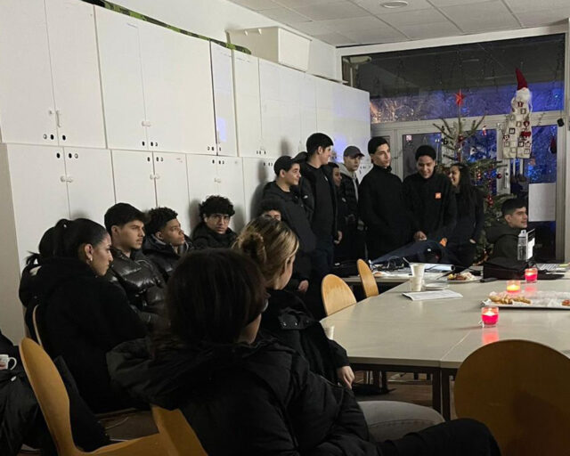 Eine Gruppe junger Menschen, meist in Schwarz gekleidet, sitzt und steht um Tische in einem mit einem Weihnachtsbaum und Kerzen geschmückten Raum. Einige sitzen, andere stehen in der Nähe von Schließfächern, und die warme Beleuchtung schafft eine gemütliche Atmosphäre.