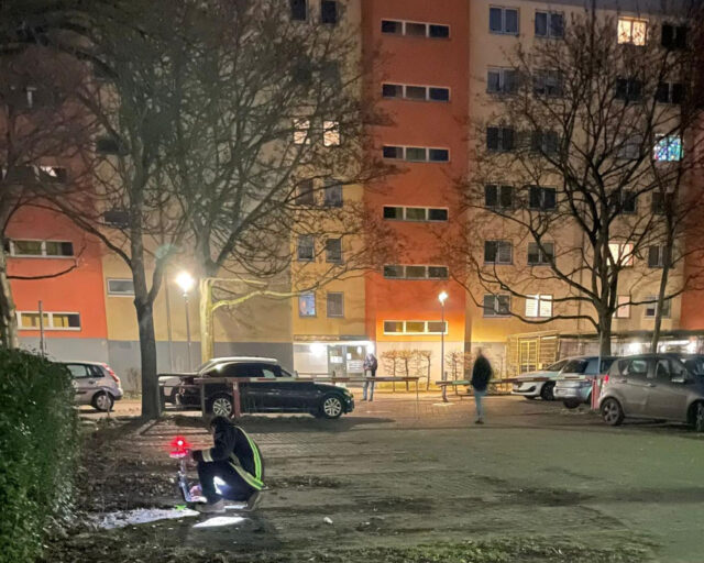 Eine Person, die eine reflektierende Weste trägt, kniet mit einer Taschenlampe auf dem Boden eines schwach beleuchteten nächtlichen Parkplatzes vor einem Wohnhaus. Im Hintergrund sind mehrere Autos und ein paar Menschen zu sehen.
