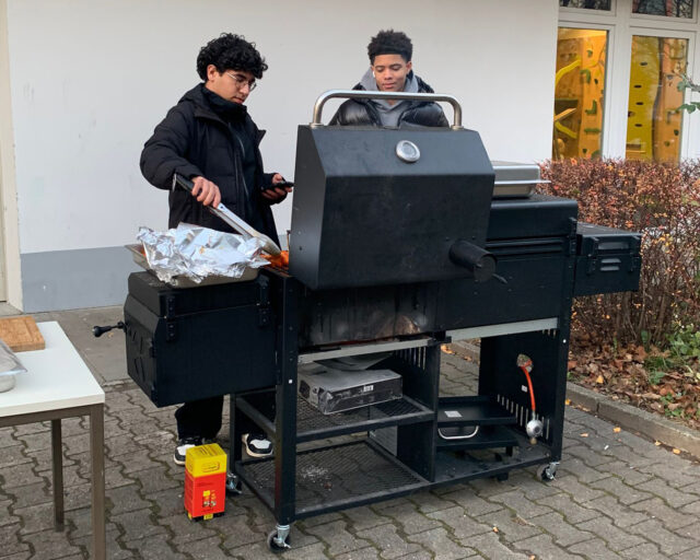 Zwei Personen stehen im Freien an einem großen schwarzen Grill. Einer wendet das Essen mit einer Zange, während der andere zusieht. Auf dem Grill und einem Tisch in der Nähe befinden sich Folienschalen und Vorräte. Im Hintergrund sind Büsche und ein Gebäude zu sehen.