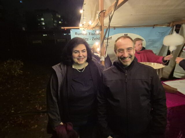 Zwei Personen, eine Frau und ein Mann, stehen lächelnd nachts im Freien unter Lichterketten in der Nähe eines Festplatzes. Hinter ihnen befinden sich Menschen und auf der rechten Seite des Bildes eine Zuckerwattemaschine.