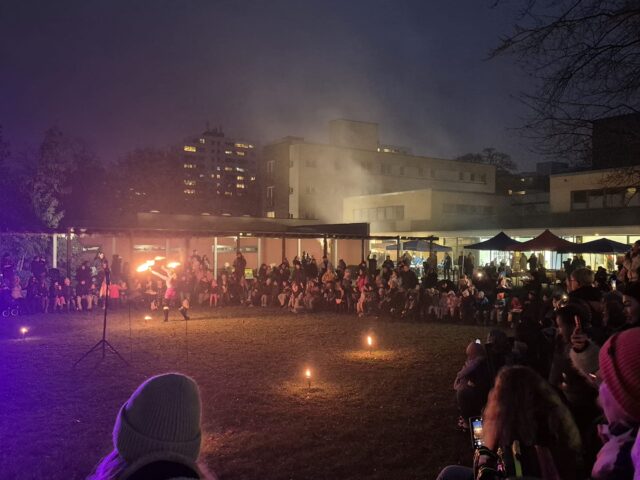 Eine Menschenmenge versammelt sich nachts im Freien und beobachtet einen Feuerkünstler in der Mitte einer Wiese. Kleine Flammen sind auf dem Boden entzündet, und im Hintergrund sind Gebäude unter dem dunklen Himmel zu sehen.
