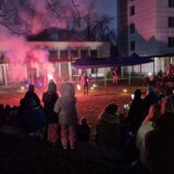 Eine Abendveranstaltung unter freiem Himmel mit einer Menschenmenge, die einem Darsteller in der Mitte zusieht, beleuchtet von bunten Lichtern und Rauch, in der Nähe von Gebäuden und Zelten, mit Menschen, die für das kalte Wetter warm angezogen sind.