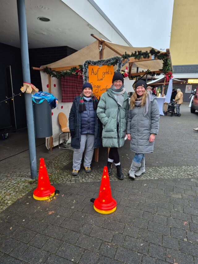Drei mit Wintermänteln und Mützen bekleidete Personen stehen lächelnd vor einem dekorierten Holzstand auf einem Markt im Freien. Zwei orangefarbene Verkehrskegel stehen vor ihnen auf dem Boden. Der Stand hat ein Schild in deutscher Sprache.