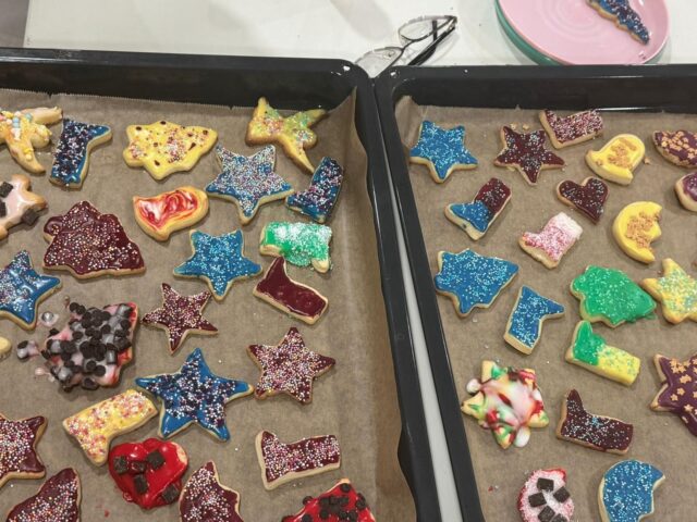 Bunte, stern- und tierförmige Kekse, die mit Zuckerguss und Streuseln verziert sind, werden auf zwei Backblechen mit Pergamentpapier angeordnet. Im Hintergrund sind ein Paar Gläser und ein rosa Teller zu sehen.