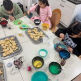 Drei Kinder sitzen an einem Tisch und verzieren Kekse mit buntem Zuckerguss und Streuseln. Verschieden geformte Kekse, Schüsseln mit Zuckerguss und Streusel sind auf dem weißen Tisch in einer Küchenumgebung ausgebreitet.