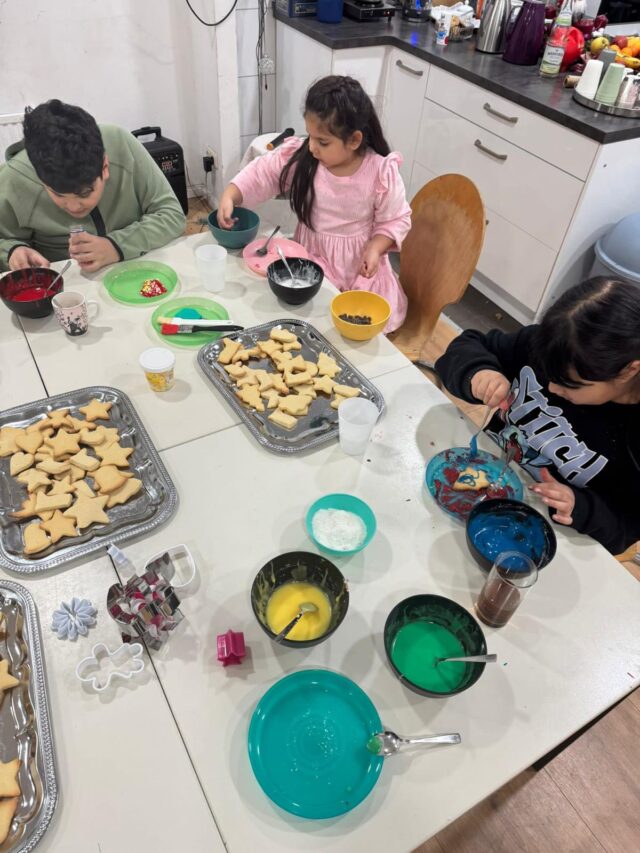 Drei Kinder sitzen an einem Tisch und verzieren Kekse mit buntem Zuckerguss und Streuseln. Verschieden geformte Kekse, Schüsseln mit Zuckerguss und Streusel sind auf dem weißen Tisch in einer Küchenumgebung ausgebreitet.