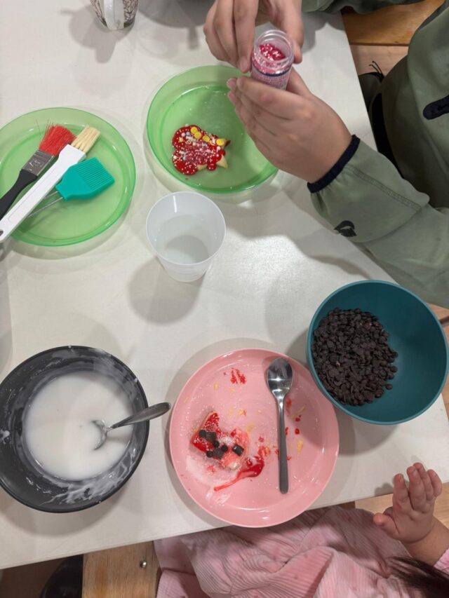 Zwei Kinder verzieren Kekse an einem Tisch. Auf den Tellern liegen ein roter Keks mit Glasur, Schokoladenstückchen, Zuckerguss in einer Schüssel und Streusel. Tassen, Löffel und Backpinsel stehen ebenfalls auf dem Tisch.