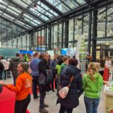 Eine Menschenmenge unterhält sich auf einer überdachten Ausstellung oder Messe mit gläsernen Decken. Einige Mitarbeiter in orangefarbenen und grünen Markenhemden sprechen mit den Besuchern. Im Hintergrund sind Stände, Banner und Informationsdisplays zu sehen.