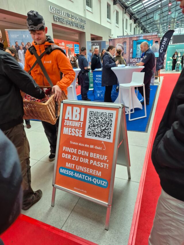 Eine Karrieremesse mit Menschen, die sich an Ständen unterhalten. Im Vordergrund steht ein Schild mit der Aufschrift "ABI Zukunft Messe" und wirbt mit einem QR-Code für ein Karrierequiz. Ein Mann in einer orangefarbenen Jacke hält einen Korb in der Hand. Der Schauplatz ist ein Innenraum.