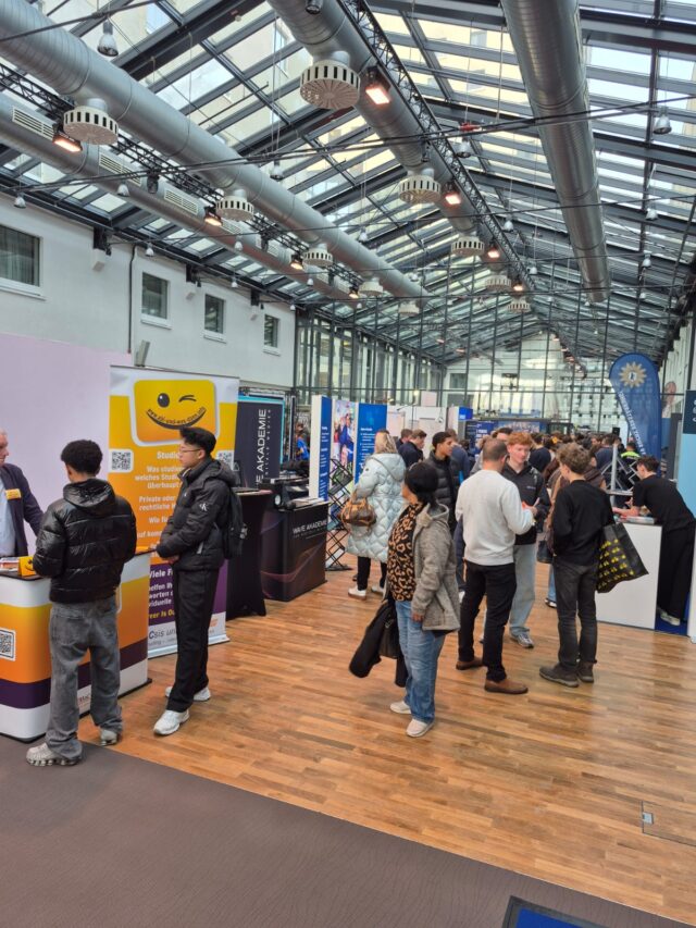 Eine Gruppe von Menschen geht umher und unterhält sich auf einer belebten überdachten Jobmesse mit verschiedenen Ständen und Bannern, die an den Seiten einer geräumigen Halle mit einer Glasdecke und einem Holzboden angebracht sind.