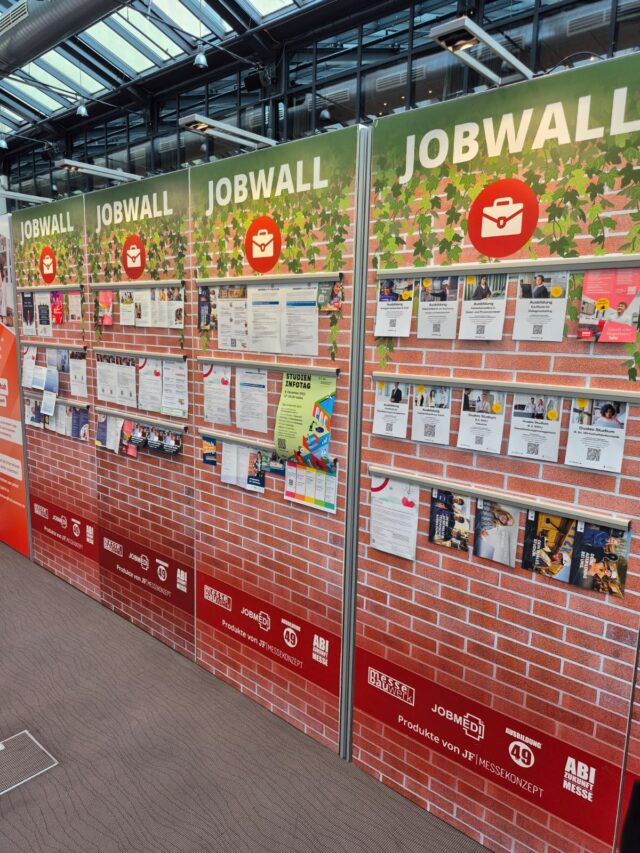 Eine Jobwand mit mehreren Paneelen, die mit Stellenausschreibungen, Flyern und QR-Codes bedeckt sind, wird in Innenräumen aufgestellt. Das Design zeigt unten ein rotes Ziegelsteinmuster und oben grünes Blattwerk.