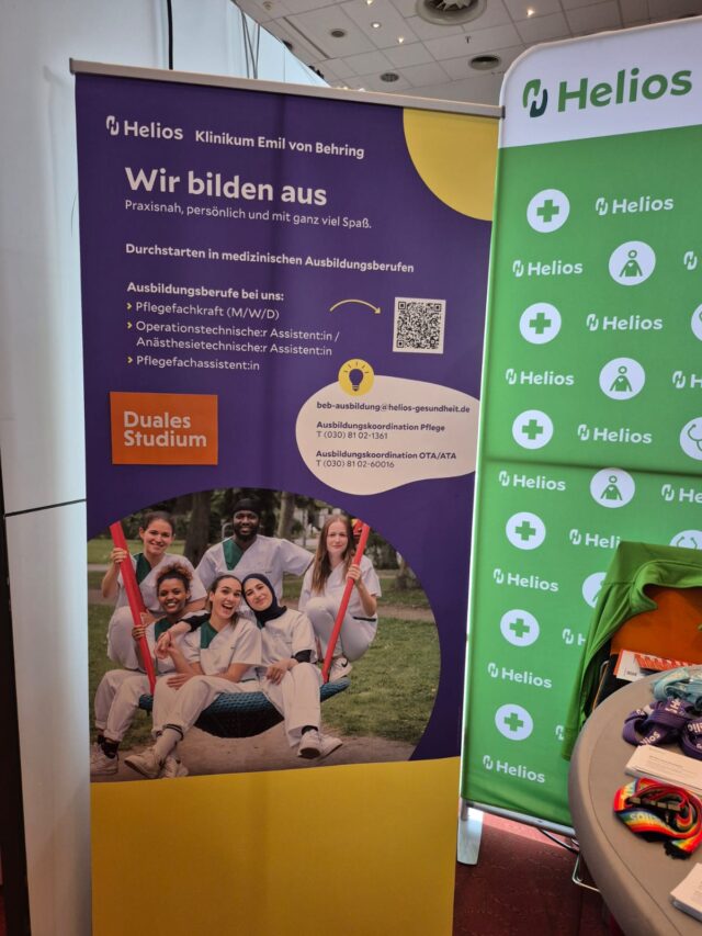 Ein Werbebanner der Helios Klinik wirbt für medizinische Ausbildungsgänge. Es zeigt sechs lächelnde junge Menschen in Kitteln, die auf einer Wiese sitzen, und informiert über duale Studienmöglichkeiten und Gesundheitsberufe. Neben dem Banner steht ein Stand mit der Marke Helios.